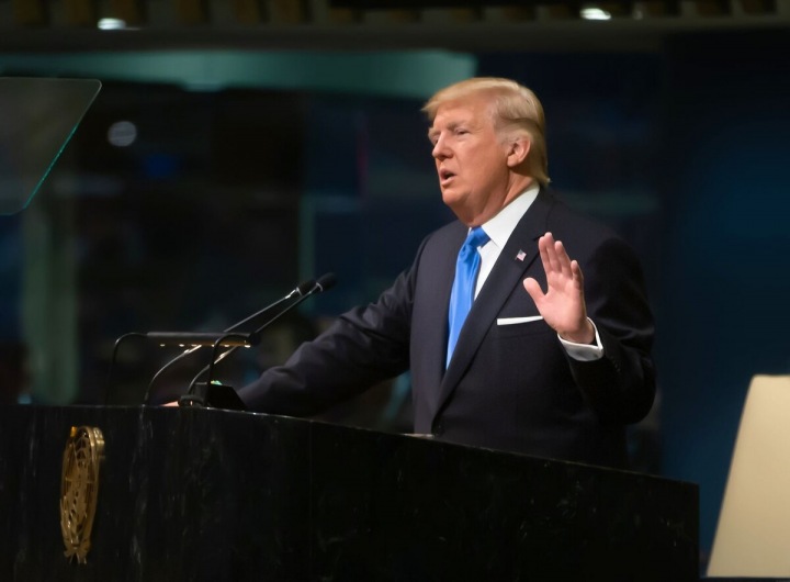 Trump stojí u řečnického pultu, na sobě má tmavý oblek a modrou kravatu. Trump stojí u řečnického pultu, na sobě má tmavý oblek a modrou kravatu.