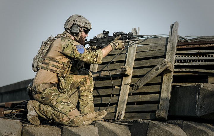 Voják s helmou, ochrannými brýlemi se zbraní v ruce za barikádou ze dřeva